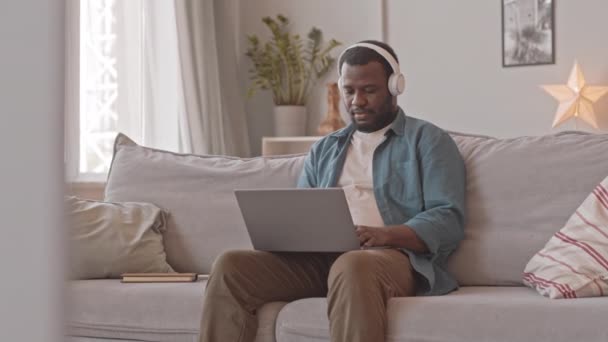 Střední Záběr Afroameričan Sluchátkách Pracuje Notebooku Sedí Pohovce Útulném Bytě — Stock video