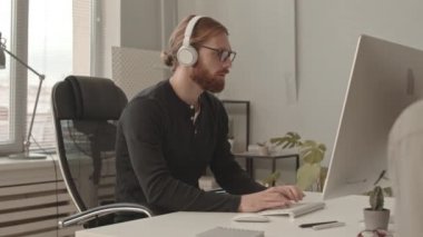 Kulaklıklı ve gözlüklü genç erkek programcının bilgisayarda çalışırken ve modern ofisteki masada oturan meslektaşıyla konuşurken ki yavaş çekimi.