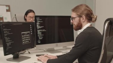 Bilgisayarda bilgisayarın program kodunu yazarak masa başında oturan bilişim uzmanının yavaş çekimde Asyalı meslektaşıyla konuşurken belden yukarı görüntüsü.