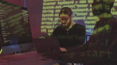 Gece vakti karanlık ofisteki bilgisayarları hackleyen iki beyaz ve Afro-Amerikalı adamın orta boy fotoğrafı. Yüzlerine yansıyan yeşil programlama kodları