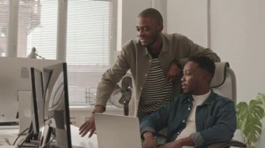 Modern ofiste çalışan bilgisayar monitörüne bakarak gündelik kıyafet giymiş iki genç Afro-Amerikalı adamın orta boy görüntüsü.