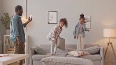 Yavaş çekimde Afro-Amerikalı bir adam akıllı telefonuyla iki tatlı kızının evlerinde yastıkların üzerinde zıplayıp gülerken videosunu kaydediyor.