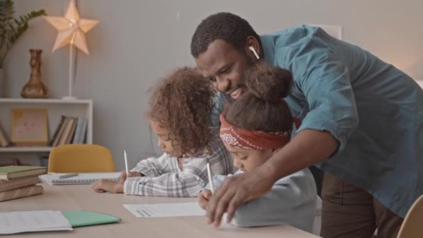 中镜头的快乐的非裔美国人帮助他的两个可爱的女儿在舒适的家里一起学习 — 图库视频影像