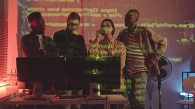 Orta yavaş çekimde, dört farklı genç programcı bilgisayar başında oturmuş sohbet ederken onların ve ofis duvarlarının üzerinde çalışan programlama kodlarıyla.