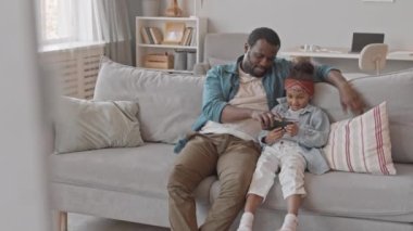 Yavaş çekimde Afro-Amerikalı bir adam ve iki tatlı kızı rahat bir evde kanepede oturup akıllı telefondan video izleyip birlikte iyi vakit geçiriyorlar.