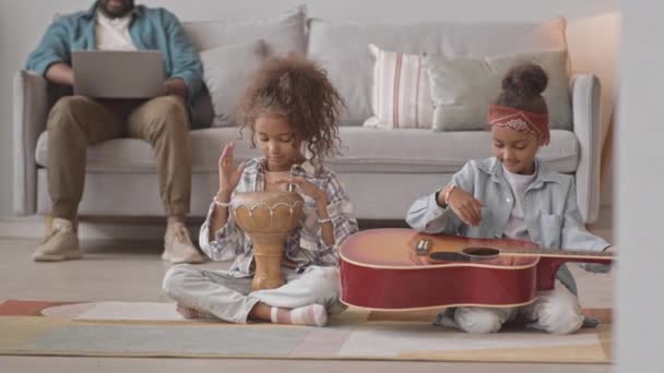 两个可爱的小女孩在客厅的地毯上弹奏吉他和非洲鼓 而她们的父亲在笔记本电脑上坐在灰色的沙发上做背景音乐 — 图库视频影像