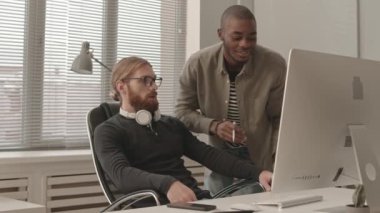 Kafkasyalı ve Afro-Amerikalı iki genç erkeğin yavaş çekimleri bilgisayar monitörüne bakıyor projeyi tartışıyorlar, modern ofiste birlikte çalışıyorlar.