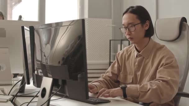 穿着便服和眼镜在现代办公室电脑上工作的年轻亚洲程序员的中档慢镜头 — 图库视频影像