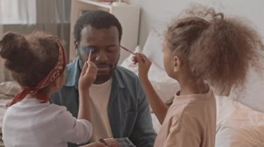 İki küçük kızı yüzüne göz farı ve diğer kozmetik ürünleri sürerken ve evde birlikte eğlenirken elleri kapalı oturan Afro-Amerikalı adamın el kamerası görüntüsü.