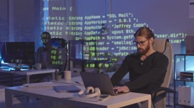 İki farklı erkek programcının iş yerindeki karanlık ofiste oturup ofis içini gösteren programlama kodlarıyla orta ölçekli görüntüleri.
