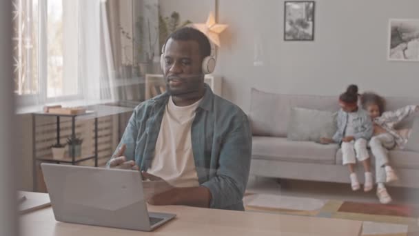 中慢镜头的非裔美国人坐在家里笔记本电脑上的耳机视频会议上 两个爱玩的女儿在后台玩乐 — 图库视频影像