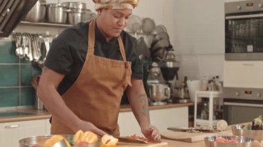 Önlük ve türban takmış, mutfak masasının yanında duran taze sebzeleri kesen ve kaseye koyan Afro-Amerikalı genç bir kadın.