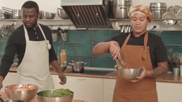 中等身材的年轻女子戴着头巾和围裙 把新鲜的沙拉和钳子混合在碗里 非洲裔美国男子则站在专业厨房的炉边做饭 — 图库视频影像