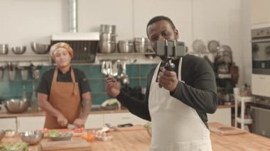 Afro-Amerikalı önlük giyen, akıllı telefonu tripoda tutan, iş yerinde video blogu çeken, sebze kesen, arka planda yemek pişiren...