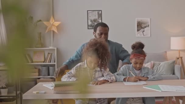 在舒适的客厅里 一个帮助女儿们做数学作业的非裔美国人坐在桌子前 拍了一张中等的照片 — 图库视频影像