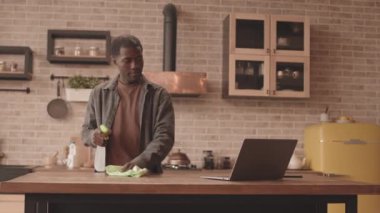 Genç Afro-Amerikalı bir adamın mutfak masasını sprey ve bezle temizlerken ve dizüstü bilgisayarında komik film ya da video izlerken modern dairesinde ev işi yaparken.