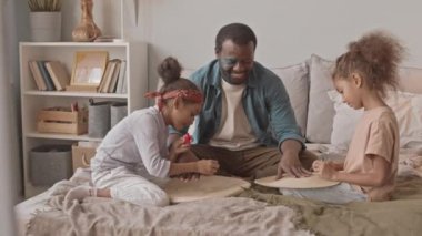 Mutlu Afro-Amerikalı bir adam ve iki küçük kızının evde birlikte eğlendikleri yavaş çekim. Kızlar babalarının tırnaklarını boyuyor.
