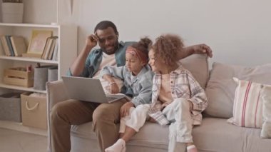 Afro-Amerikalı bir adam ve iki güzel kızının, rahat bir dairede gri koltukta oturup dizüstü bilgisayarlarında komik videolar izledikleri bir fotoğraf.