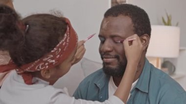 İki küçük kızı yüzüne komik bir makyaj yaparken gözleri kapalı oturan, gülümseyen Afro-Amerikan bir adamın orta boy yakın çekimi.