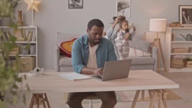 Mikrofonda şarkı söyleyen iki küçük kızın laptopta babalarını rahatsız ederken orta boy görüntüsü. Afro-Amerikalı adam, çocukların sesini duymamak için kulaklık takıyor.