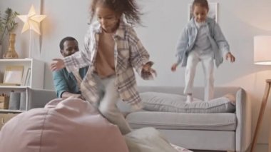 Afro-Amerikalı babaları akıllı telefon kamerasıyla oynarlarken iki küçük kız kardeşin rahat evlerinde yastıkların üzerinde zıplayıp eğlenirken ki yavaş çekimleri.