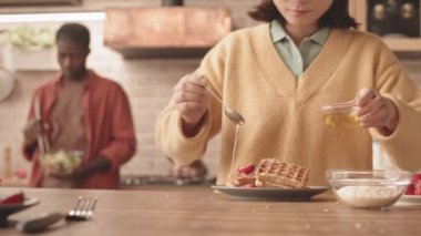 Afrikalı Amerikalı erkek arkadaşı arka planda salatayı karıştırırken, çilekli waffle 'ları bal ve hindistan ceviziyle süslerken, Asyalı genç bir kadının yavaş çekimleri.