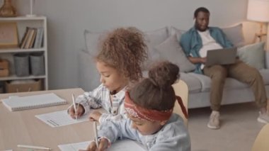 Afro-Amerikalı babaları arka planda dizüstü bilgisayarda otururken, iki küçük kız kardeşin rahat oturma odasında oturup matematik sınavı yaptığı yavaş çekim.
