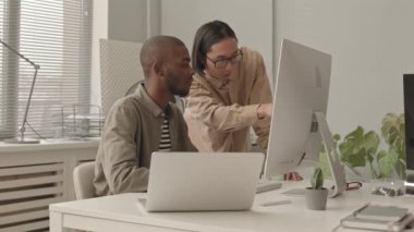 İki Afro-Amerikalı ve Asyalı programcının modern ofiste bilgisayar üzerinde çalışan yazılımları tartışırken orta yavaş çekimleri.