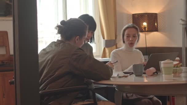 大学のテーブルで一緒に座って宿題をしている女性のクラスメートと話す障害のある男性大学生の中低速ショット — ストック動画