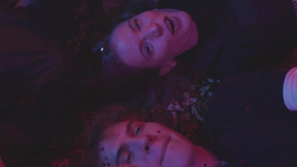 上から20代の多様な友人のグループのスロモショットを一緒にカメラを見て 紫色の照明でネオンパーティーで踊るコンフェッティの床に横たわっている — ストック動画
