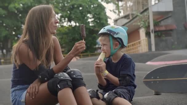 一位活跃的妈妈和7岁的儿子膝部和肘部垫坐在露天溜冰场的地面上 一边吃着冰激凌 一边聊天 — 图库视频影像