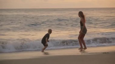 Genç, güzel bir kadın ve 7 yaşındaki oğlu sahilde günbatımının tadını çıkarıyorlar, dalgalardan kaçıyorlar ve gülüyorlar.