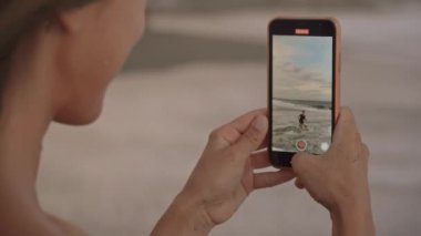 Küçük oğlunun gün batımında denizde oynarken çekilmiş akıllı telefonuyla gülümseyen güzel bir kadının orta boy yakın çekimi.