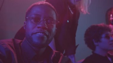 Gece kulübünde neon ışıklandırmalı çeşitli arkadaşlarla parti yaparken moda gözlüklü, kameraya konuşan Afro-Amerikan bir adamın yavaş çekimleri.