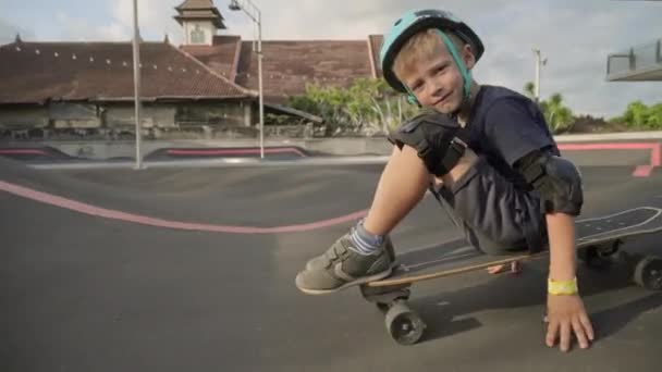 ヘルメットと膝と肘パッドでかわいい7歳の白人の男の子の追跡ショットスケートボードに座って転がりスケートパークでカメラに笑顔 — ストック動画