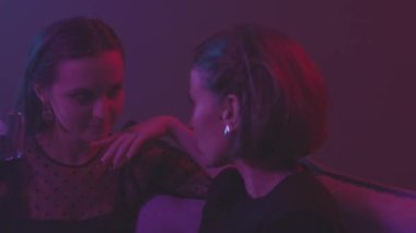 İki güzel beyaz kadının sohbet edip şarap içtiği, neon renkli bir partide mor ışıklı koltukta oturduğu ağır çekim.