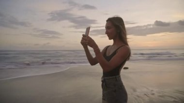 Genç ve güzel bir kadın, akşamları sahilde duran akıllı telefonuyla inanılmaz günbatımının fotoğraflarını çekiyor.