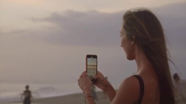 Akıllı telefonlu genç ve çekici bir kadının kumsalda gün batımının güzel resimlerini çekerken.
