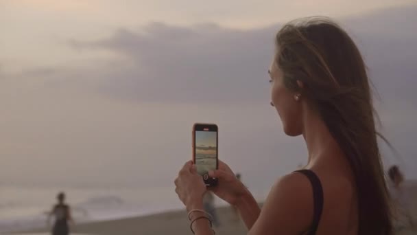 弧形的年轻迷人女子与智能手机在海滩拍摄美丽落日的照片 — 图库视频影像