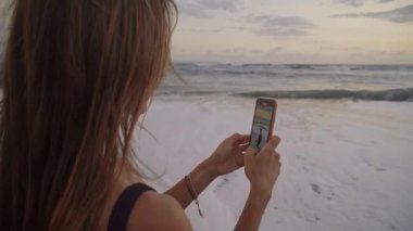 Genç ve güzel bir kadının, oğlunun gün batımında deniz kıyısında yürüyüşünün akıllı telefonuyla video kaydını çekerken.