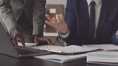 Düşük açılı tanınmayan erkek yönetici masasında oturuyor, proje belgeleriyle çalışıyor, gündüzleri ofisteki taşınabilir bilgisayarı kullanıyor.