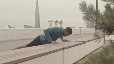 Yavaş çekimde zayıf, Afro-Amerikan bir adam soğuk bir sabahta briçte yedek kulübesinde şınav çekiyor.