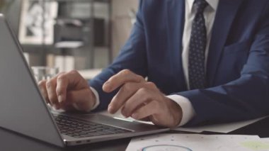 Mavi resmi kıyafetler giyen erkek CEO 'nun düşük açılı elleri ofiste oturuyor, dizüstü bilgisayarında daktilo yazıyor.