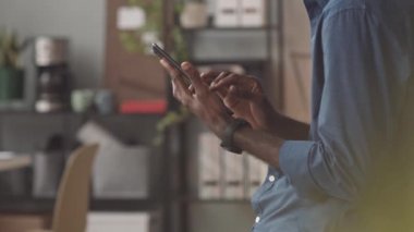 Kırpılmış siyahi adamın gündüz vakti evde dikilip akıllı telefon kullanarak saatine bakarken alçak açılı yan görüntüsü.
