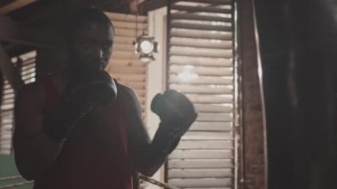 Boks eldivenli Afrikalı-Amerikan boksörün boks antrenmanı sırasında kum torbasına yumruk attığı orta yavaş bir kare.