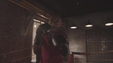 Elde tutulan orta boy pozlar, boks eldivenli kaslı siyahi bir adamın ringlere boks yumrukları atarken kameraya öfkeyle bakması.