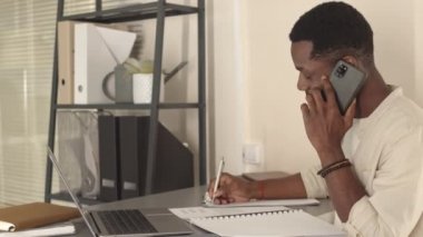 İş günü masa başında oturan, akıllı telefondan konuşan ve not alan Afrikalı Amerikalı genç bir adamın belden yukarı görüntüsü.