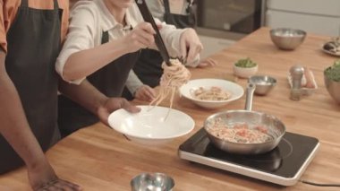 Kafkasyalı genç bir kadının maşa ve kepçe kullanarak tavadan makarnayı tabağa koyması. Afro-amerikan erkek eli, Asyalı aşçılık öğrencisi...