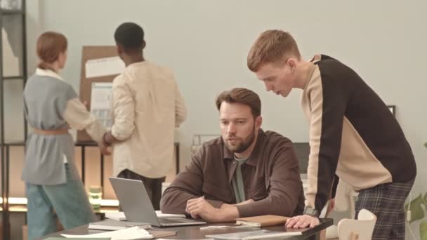 中くらいの長さの髭を生やした白人マネージャーが机の上に座ってノートパソコンを使って近くに立って話している若い男性従業員2人の同僚が — ストック動画