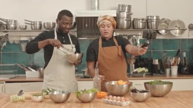 Sarık ve önlük takan genç bir kadın ve profesyonel mutfakta masada oturan Afro-Amerikan bir adam tripodun üzerindeki akıllı telefon kamerasında yemek ve malzemelerini gösteriyor.